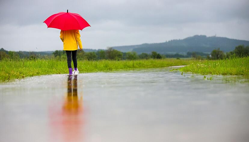 Eine Spazierg&auml;ngerin in Gummistiefeln l&auml;uft im Regen auf einem &uuml;berfluteten Weg entlang.