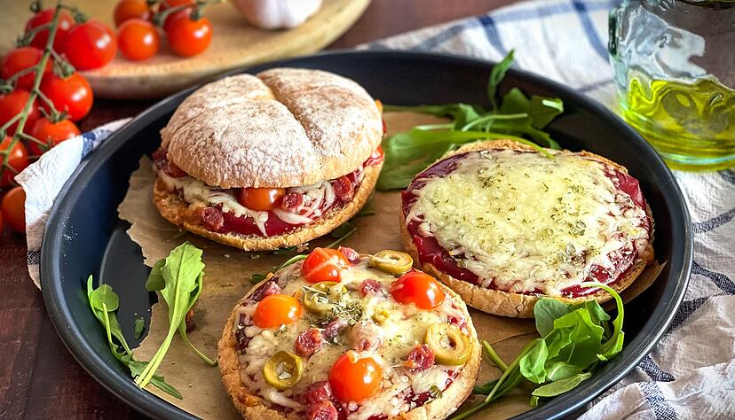 Ein rustikales Hamburgerbr&ouml;tchen wird zur Pizzabasis, belegt mit Tomatenketchup, geschmolzenem Mozzarella und mediterranen Zutaten.