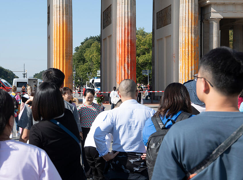 Touristen stehen vor dem Brandenburger Tor, das Mitglieder der Klimaschutzgruppe Letzte Generation am Sonntagvormittag mit oranger Farbe angespr&uuml;ht haben.