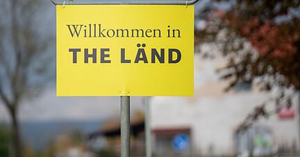 An einem Ortsschild hängt ein Zusatzschild mit der Aufschrift «Willkommen in THE LÄND».