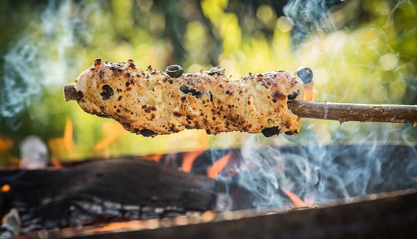 Ein Oliven-Feta-Knoblauch-Hefeteig ist um einen Haselnuss-Stock gewickelt und wird über dem Lagerfeuer gebacken.