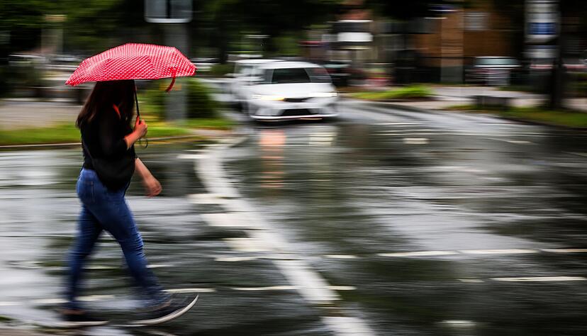 Eine Frau mit einem Regenschirm &uuml;berquert im Regen eine Stra&szlig;e. Nach grauen und verregneten Tagen sehnen viele Menschen die Sonne herbei.