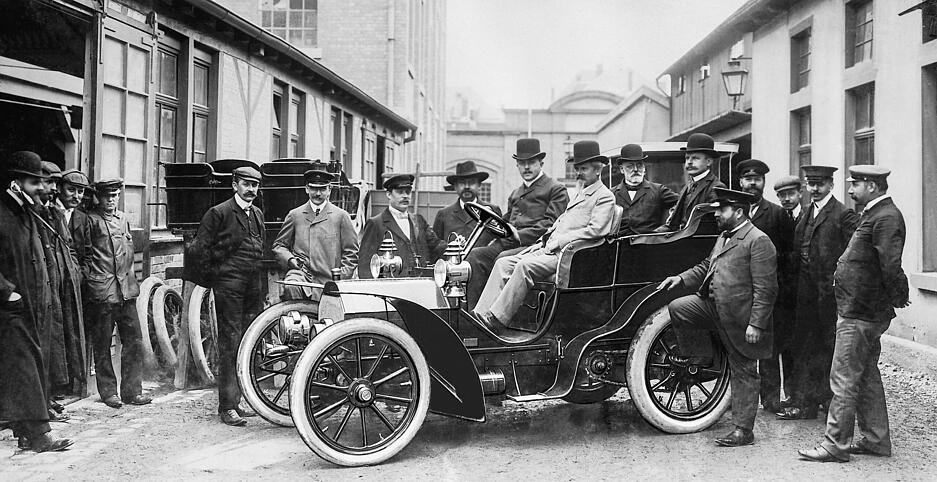 Wilhelm Maybach 1902 am Steuer eines Mercedes Simplex Wagen in der Cannstatter Fabrik der Daimler-Motoren-Gesellschaft. Im Fahrzeug hinten rechts: Adolf Daimler, Sohn von Gottlieb Daimler. Der Namenszusatz „Simplex“ sollte auf die vereinfachte Bedienbarkeit hinweisen.