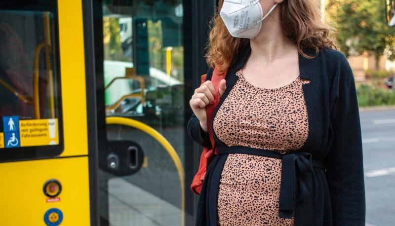 Eine schwangere Frau wartet mit Mund-Nasen-Schutz an einer Haltestelle auf dem Bus.