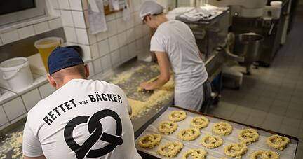Ein Bäcker und eine Auszubildende stehen am frühen Morgen mit einem T-Shirt „Rettet uns Bäcker“ in der Backstube der Bäckerei Fahrenhorst in Hannover.