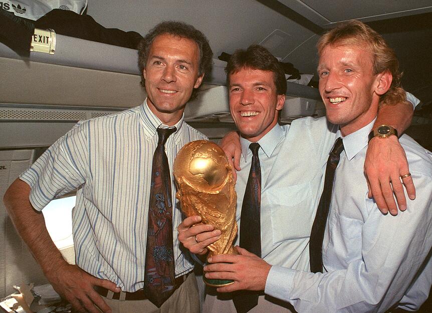 Im Flugzeug beim Rückflug von Rom nach Frankfurt präsentieren der damalige DFB-Teamchef Franz Beckenbauer (l), Kapitän und Mittelfeldspieler Lothar Matthäus (M) und Abwehrspieler Andreas Brehme, der das entscheidende Tor schoss, den gewonnenen WM-Pokal.