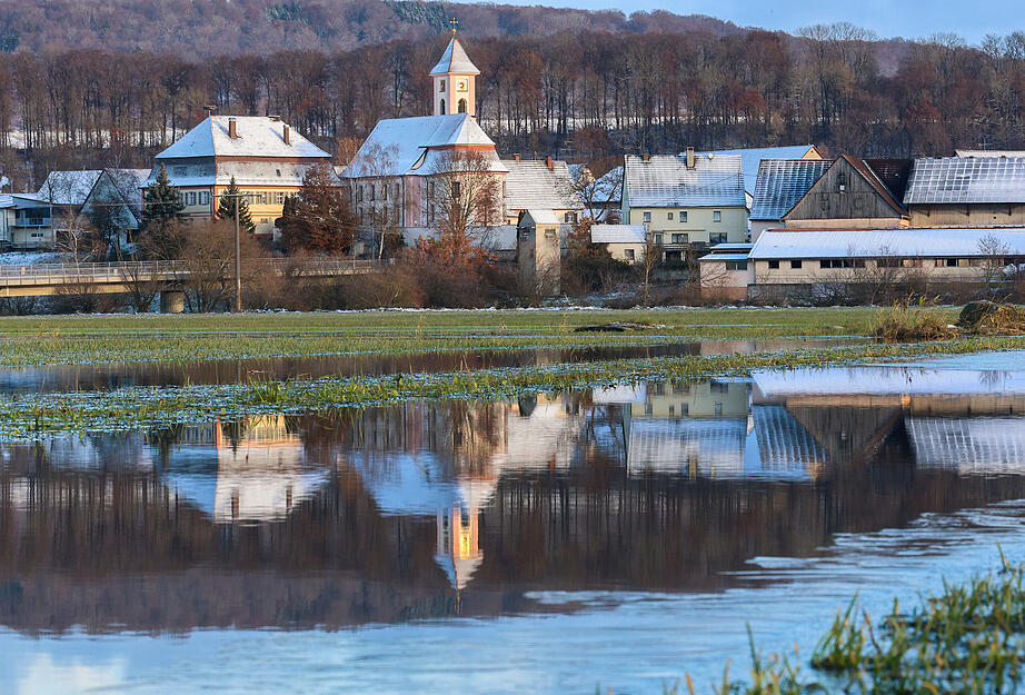 Die mit Schnee bedeckte Kirche St. Gallus in Riedlingen Zell spiegelt sich in einer überfluteten Wiese wieder.