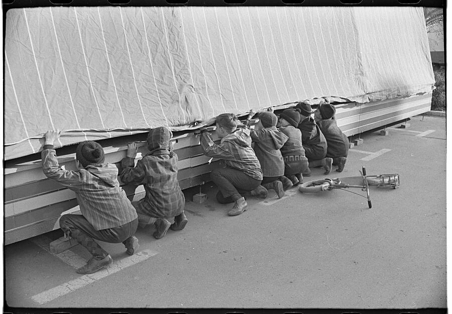 Neugierige Kinder gucken unter die Plane des Boxauto-Fahrgeschäfts zum Heilbronner Pferdemarkt im Jahr 1964.
