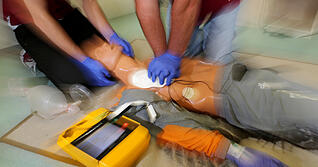 Zwei Männer demonstrieren eine Reanimation an einer Puppe, bei der ein Defibrillator zum Einsatz kommt.