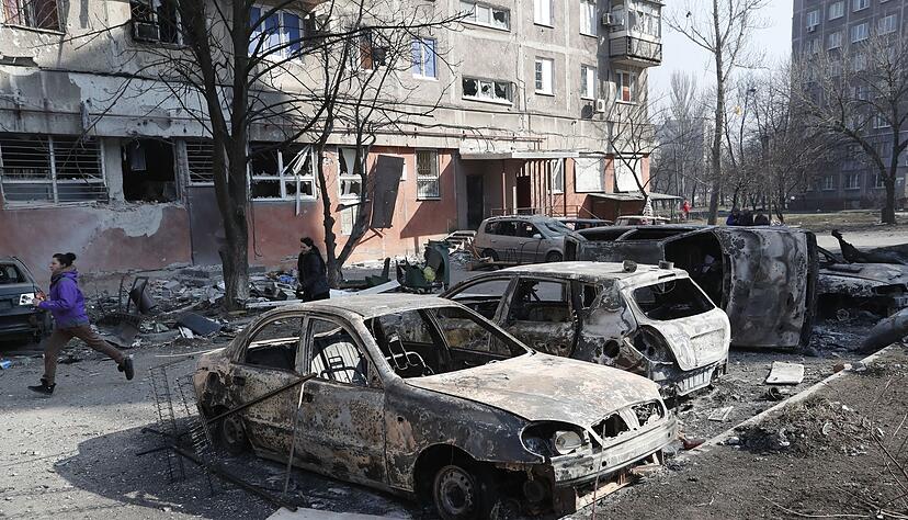 Menschen gehen an zerstörten Autos und Trümmern in Mariupol vorbei. Menschen gehen an zerstörten Autos und Trümmern in Mariupol vorbei.