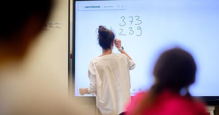 Eine Lehrerin schreibt eine Mathematikaufgabe auf eine digitale Schultafel im Klassenraum einer 4. Klasse einer Grundschule.