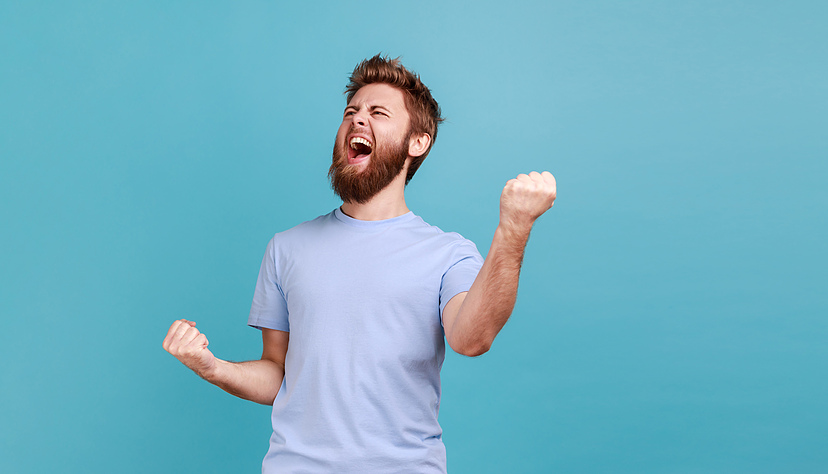 Portrait of overjoyed bearded man standing with excited expression, raising fists, screaming, shouting yeah, celebrating his victory, success. Indoor studio shot isolated on blue background.