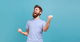 Portrait of overjoyed bearded man standing with excited expression, raising fists, screaming, shouting yeah, celebrating his victory, success. Indoor studio shot isolated on blue background.