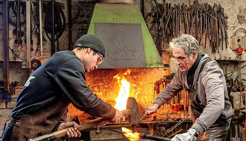 Josha Wewoda haut mit einem gro&szlig;en Hammer auf den Schrotmeisel in der Hand seines Vaters Martial Herbst. Dieser dreht das zu einem Nagel vorgeformte St&uuml;ck mit der gl&uuml;henden Schnittkante, so dass dieser Teil vom Rohmaterial getrennt werden kann. Die Ledersch&uuml;rzen sch&uuml;tzen die beiden vor Hitze und Funken.
Fotos: Christiane Kunz