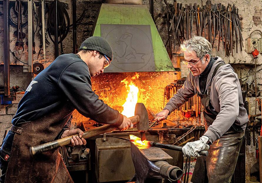 Josha Wewoda haut mit einem gro&szlig;en Hammer auf den Schrotmeisel in der Hand seines Vaters Martial Herbst. Dieser dreht das zu einem Nagel vorgeformte St&uuml;ck mit der gl&uuml;henden Schnittkante, so dass dieser Teil vom Rohmaterial getrennt werden kann. Die Ledersch&uuml;rzen sch&uuml;tzen die beiden vor Hitze und Funken.
Fotos: Christiane Kunz
