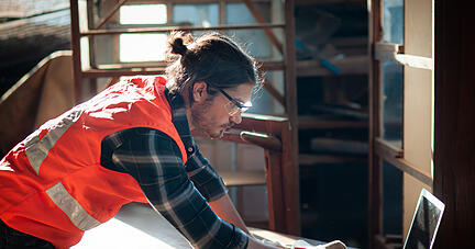 Young and handsome caucasian carpenter in the carpentry shop is working and checking blueprint in laptop to repair a furniture.