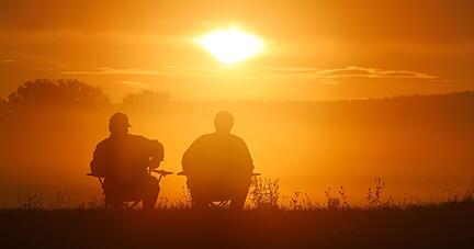Zwei Angler sitzen auf Campingstühlen und schauen sich den Sonnenaufgang in Baden-Württemberg an.