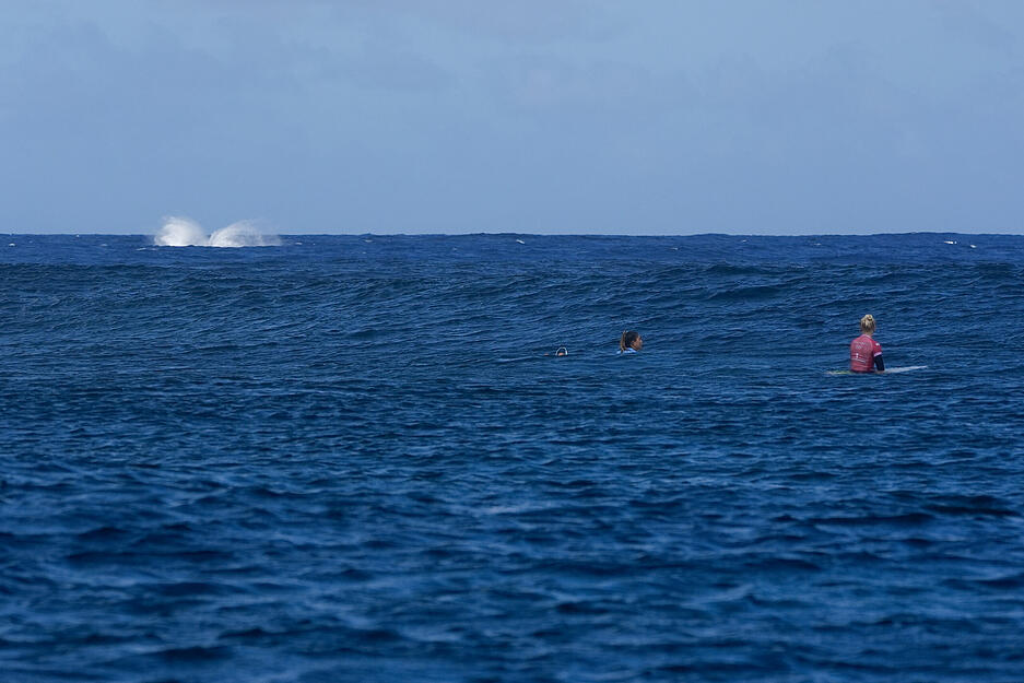 Ein Wal landet nach einem Sprung im Wasser, während Brisa Hennessy aus Costa Rica und Tatiana Weston-Webb aus Brasilien auf die nächste Welle warten.