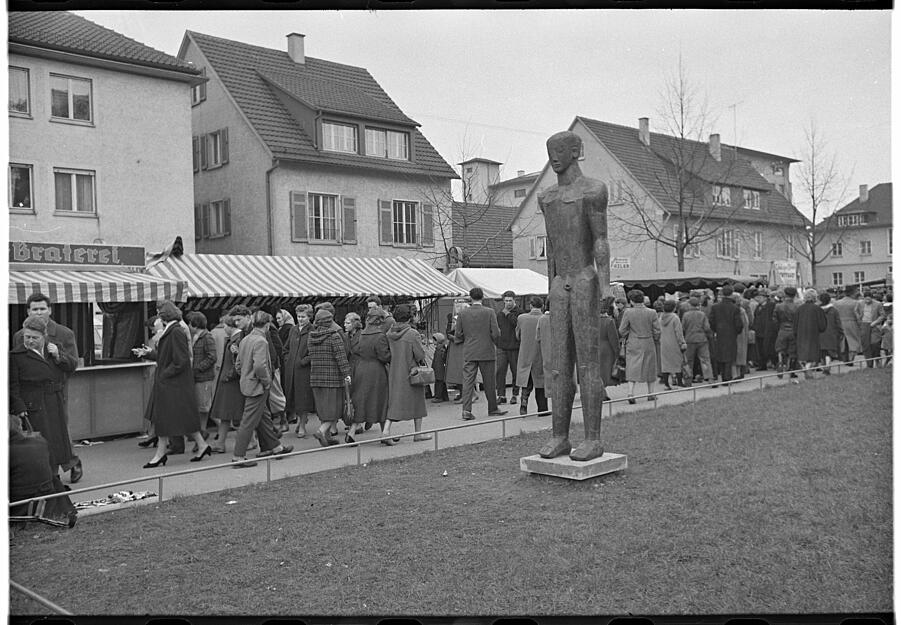 Besucher des Pferdemarkts 1959 spazieren entlang der Buden. Rechts steht die Skulptur „Der Jüngling“ von Gottfried
Gruner.
