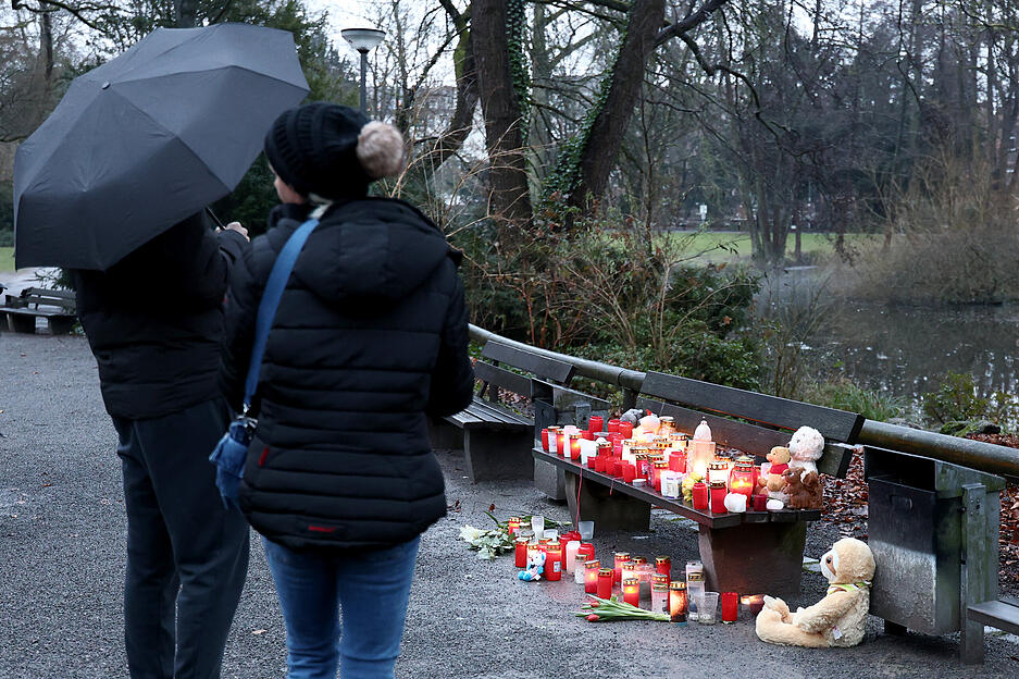 Personen stehen nach dem tödlichen Angriff in einem Park am Tatort vor einer Bank mit Trauerkerzen, Blumen und Kuscheltieren.
