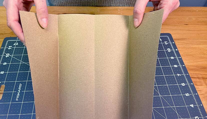Die kurze Seite des Kraftpapiers mittig nach oben falten und anschließend wieder aufklappen. Nun die Enden jeweils bis zur Falz klappen, sodass vier gleich breite Abschnitte entstehen. Die kurze Seite des Kraftpapiers mittig nach oben falten und anschließend wieder aufklappen. Nun die Enden jeweils bis zur Falz klappen, sodass vier gleich breite Abschnitte entstehen.