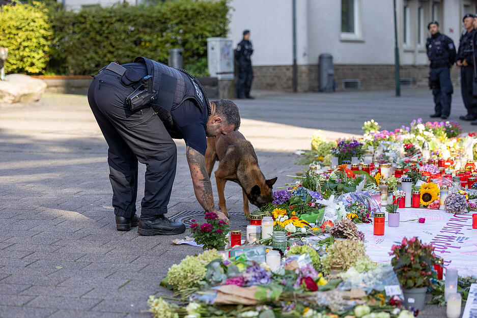 Ein Spürhund der Polizei kontrolliert vor dem Besuch des Bundeskanzlers Scholz einen Gullideckel und die zum Gedenken der Opfer abgelegten Blumen.