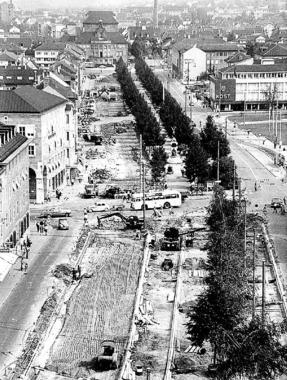 Umbau 1959: Für die künftige Hauptverkehrsstraße wird der Grünstreifen in der Mitte der Allee auf etwa ein Drittel verschmälert. Die Umrisse sind vorn in der Bildmitte erkennbar. Der Raumgewinn wird auf Gehwege und mehr Fahrspuren verteilt. Oben bildet das Theater den nördlichen Abschluss der Allee.