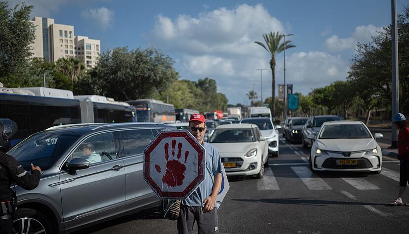 Ein israelischer Demonstrant geht mit einem Schild auf einer Stra&szlig;e, w&auml;hrend Autos sich stauen.