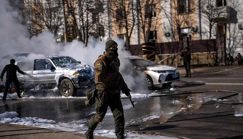 Ein ukrainischer Soldat bezieht Stellung vor einer Militäreinrichtung, während Feuerwehrleute im Hintergrund zwei brennende Autos löschen. Ein ukrainischer Soldat bezieht Stellung vor einer Militäreinrichtung, während Feuerwehrleute im Hintergrund zwei brennende Autos löschen.