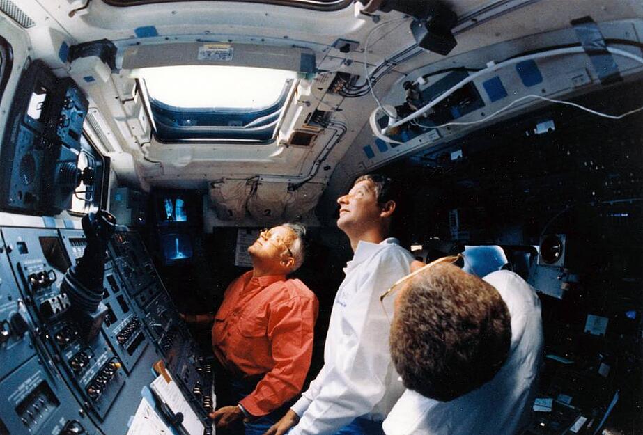 Drei STS-31 Crew-Mitglieder schauen am 25. April 1990 durch das Fenster des Space-Shuttles Discovery zum Weltraumteleskop Hubble. Bruce McCandless ist ganz links zu sehen.