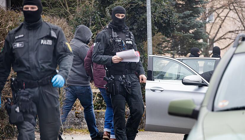 Hier wird der Beschuldigte, mittlerweile in grauer Kapuzenjacke und Jeans, in das Einsatzfahrzeug gebracht und weggefahren. Abgeschirmt ist er von vermummten Beamten.