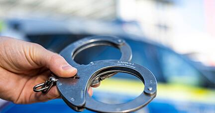 Eine Hand hält Handschellen vor einen Streifenwagen der Polizei. Eine Hand hält Handschellen vor einen Streifenwagen der Polizei.