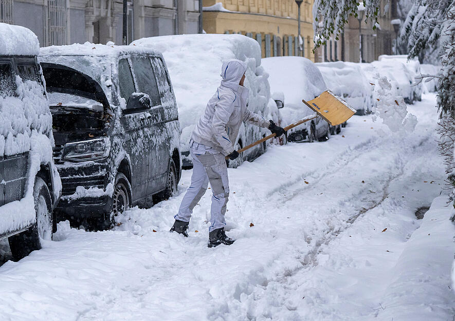 Eine Frau schippt Schnee neben ihrem eingeschneiten parkenden Auto.