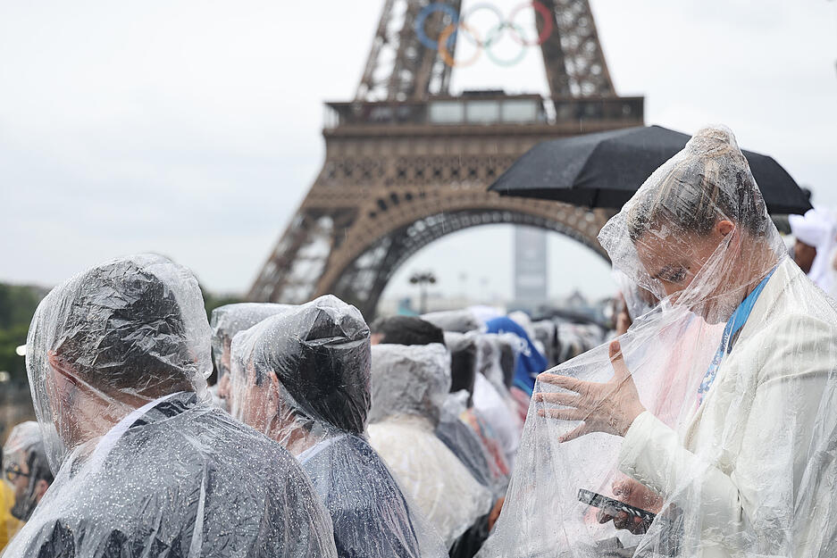 Zuschauer sitzen mit Regenschutz bedeckt auf den Tribünen vor dem Eiffelturm.