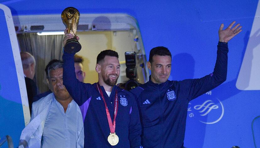 Lionel Messi (l) h&auml;lt den Weltmeisterpokal in der Hand, w&auml;hrend er mit seinem Trainer Lionel Scaloni aus dem Flugzeug aussteigt.