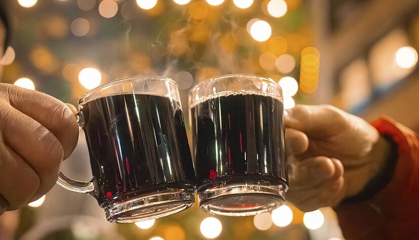 Zwei Hände halten Glühweintassen in einer festlichen Nacht mit Lichtern. (Symbolbild)